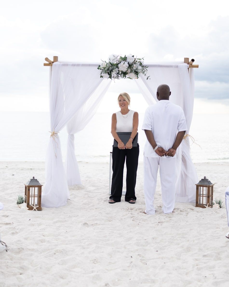 Officiant Beth Schell smiles warmly beneath a white draped floral arch on the beach, holding her ceremony folder, the Gulf of Mexico behind her.