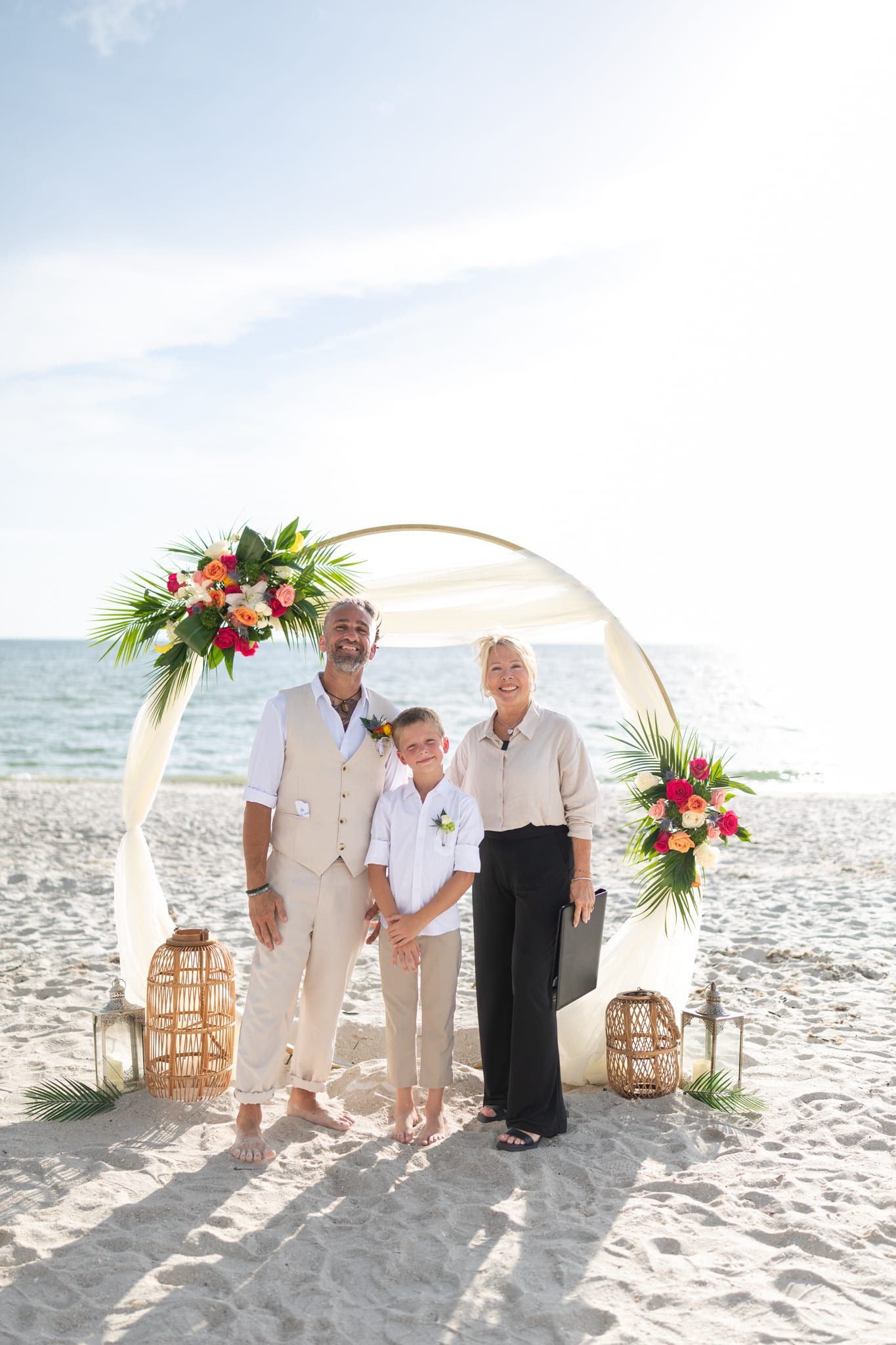 Officiant Beth Schell stands smiling with a groom and his son beneath a circular floral arch on the beach after the ceremony.