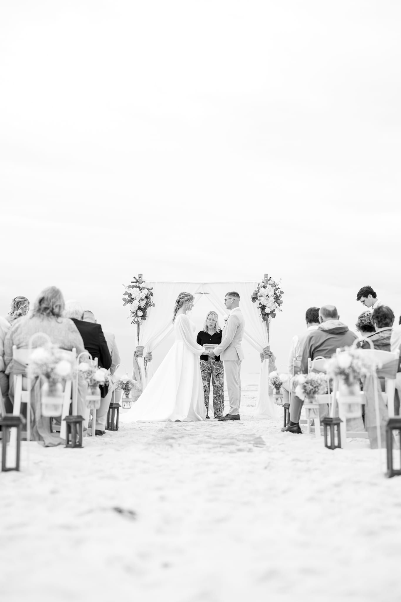 A dreamy black and white view down the sandy aisle as a bride and groom hold hands before officiant Beth Schell under a draped arch, guests seated on both sides.