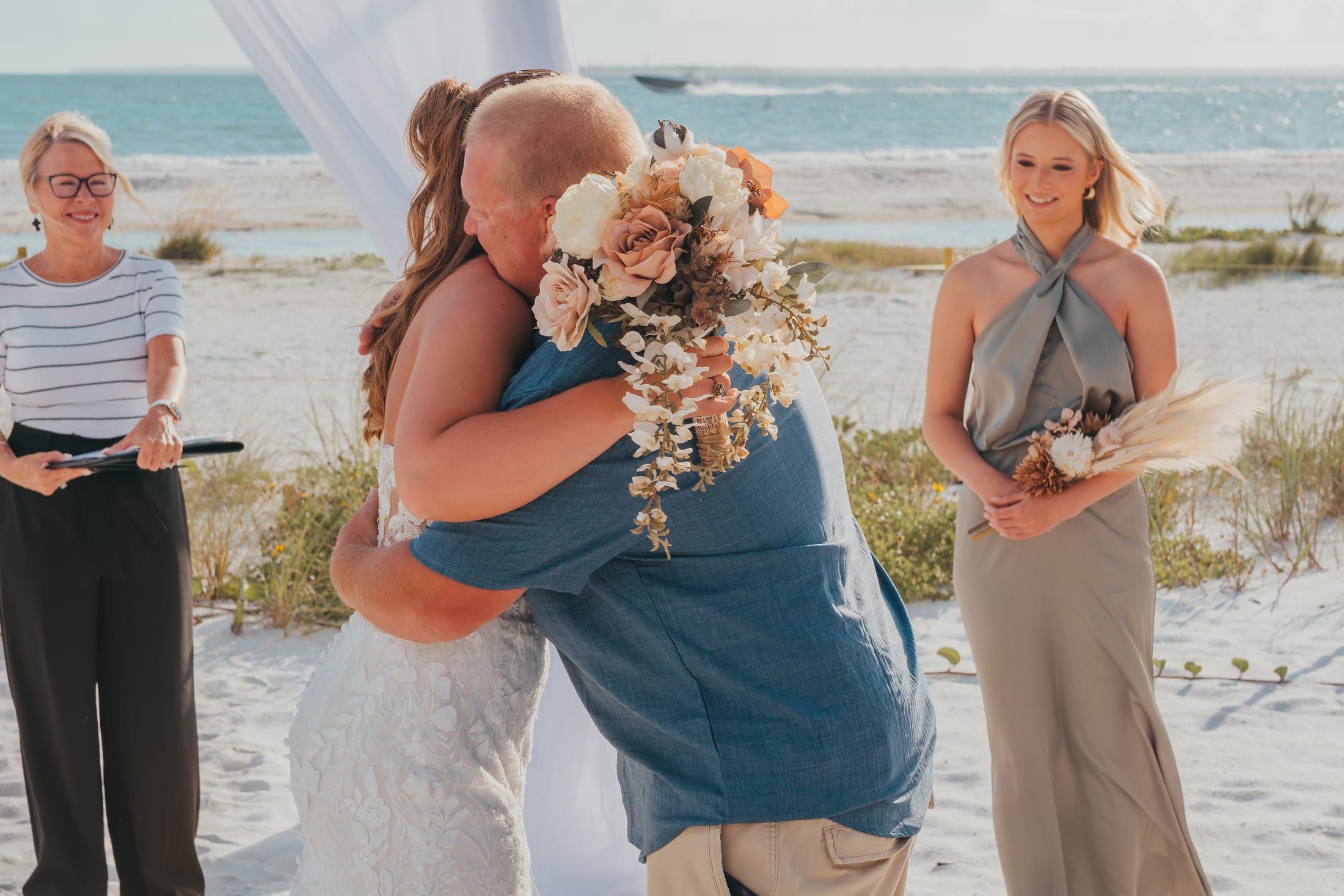 A bride is wrapped in a joyful embrace at the end of the aisle, her bouquet in hand, as officiant Beth Schell smiles nearby.