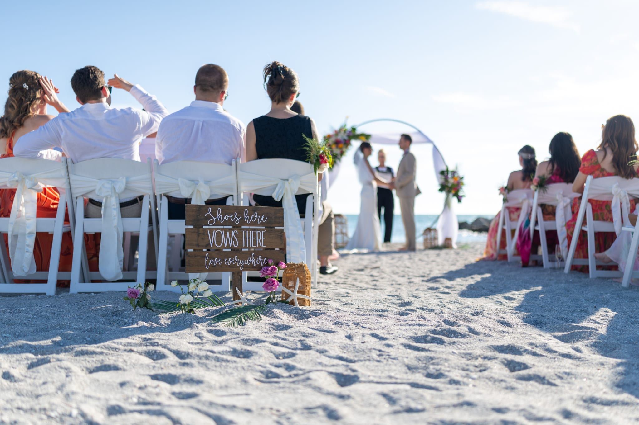 A wooden sign reading Shoes here, Vows there, love everywhere stands in the sandy aisle as guests watch a beach ceremony led by officiant Beth Schell.