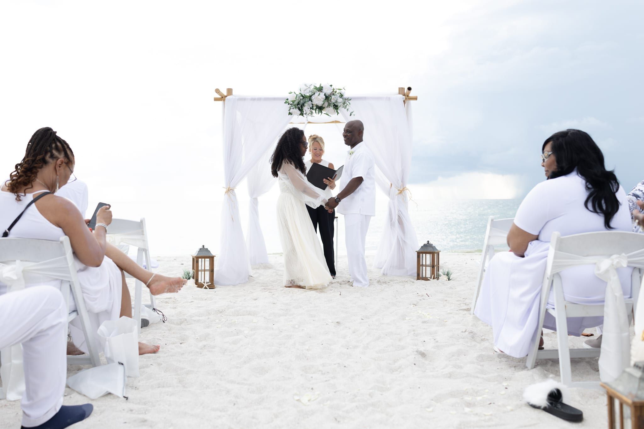 Officiant Beth Schell reads from her folder as a couple dressed in white holds hands beneath a white canopy arch on the beach, guests in white seated around them.