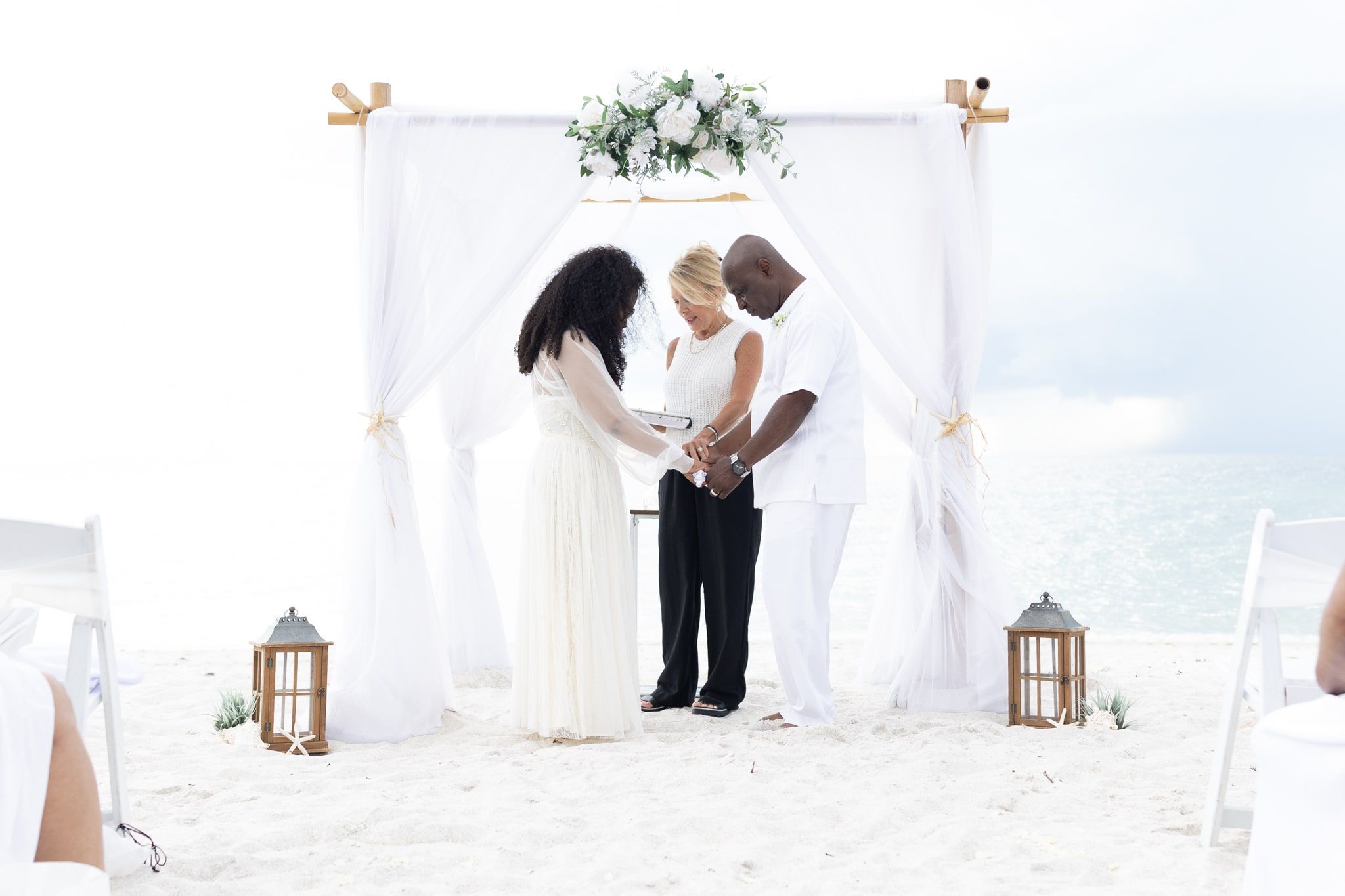 A bride and groom hold both hands under a white floral arch as officiant Beth Schell leads their vows beside the turquoise Gulf of Mexico.