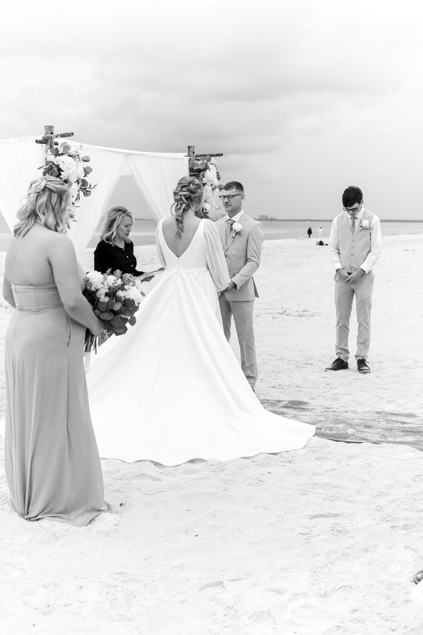 A bride and groom hold hands under a draped bamboo arch as officiant Beth Schell reads their vows, in a black and white beach photograph.