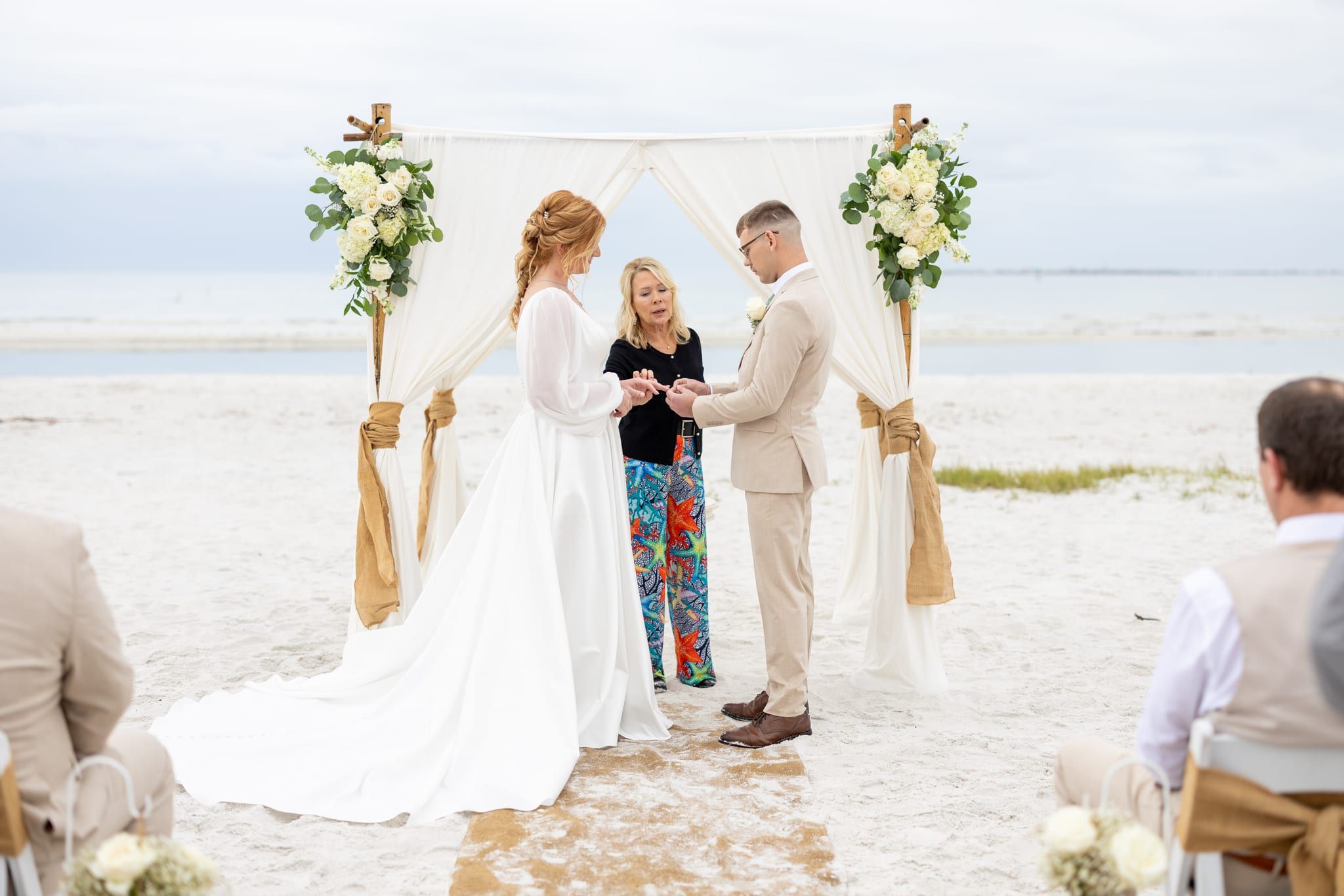 A bride and groom hold hands under a draped bamboo arch as officiant Beth Schell reads their vows beside the Gulf.