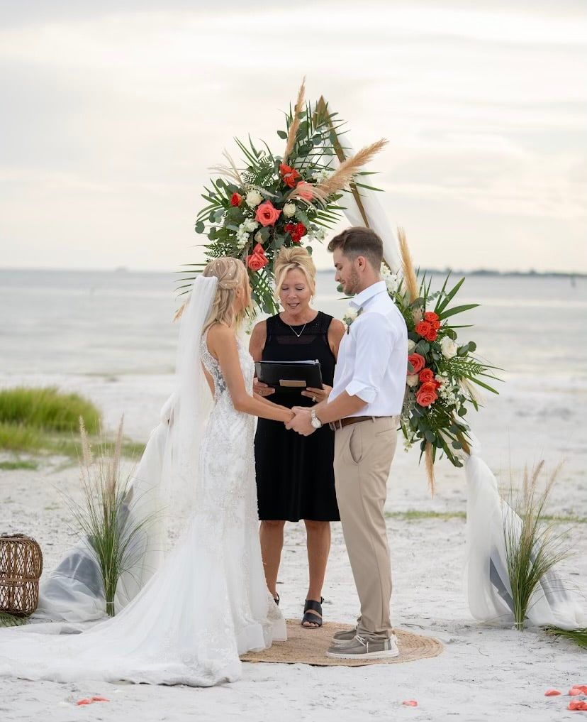 A bride and groom clasp hands under a boho pampas grass and rose arch on the beach while officiant Beth Schell reads the ceremony.