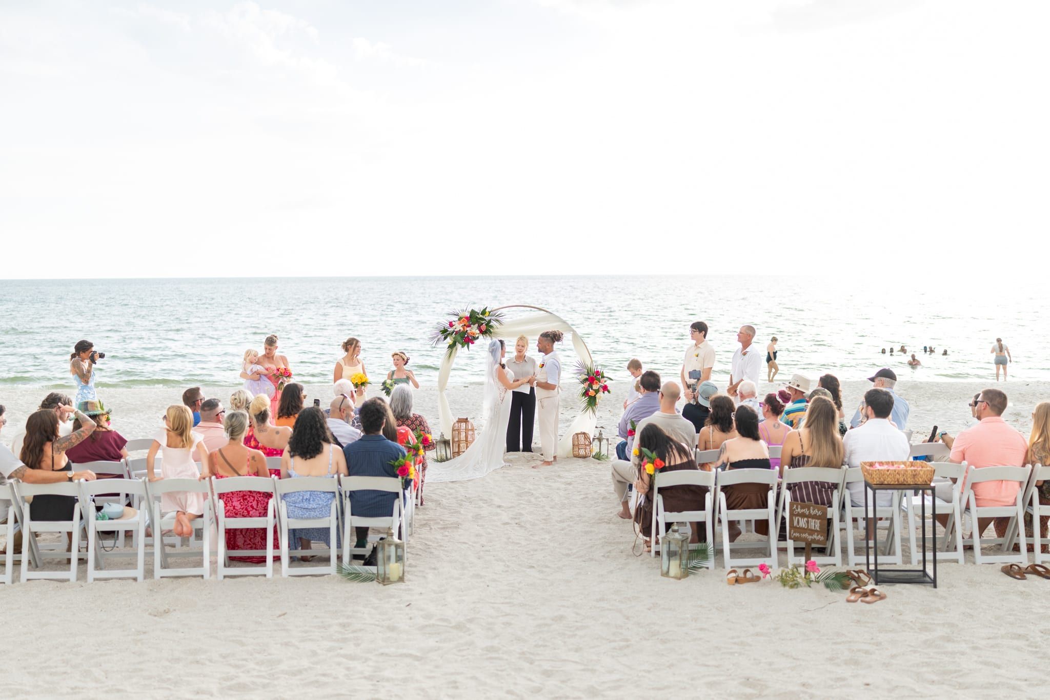 A full beach wedding with rows of seated guests watching a couple exchange vows with officiant Beth Schell under a circular arch by the Gulf.