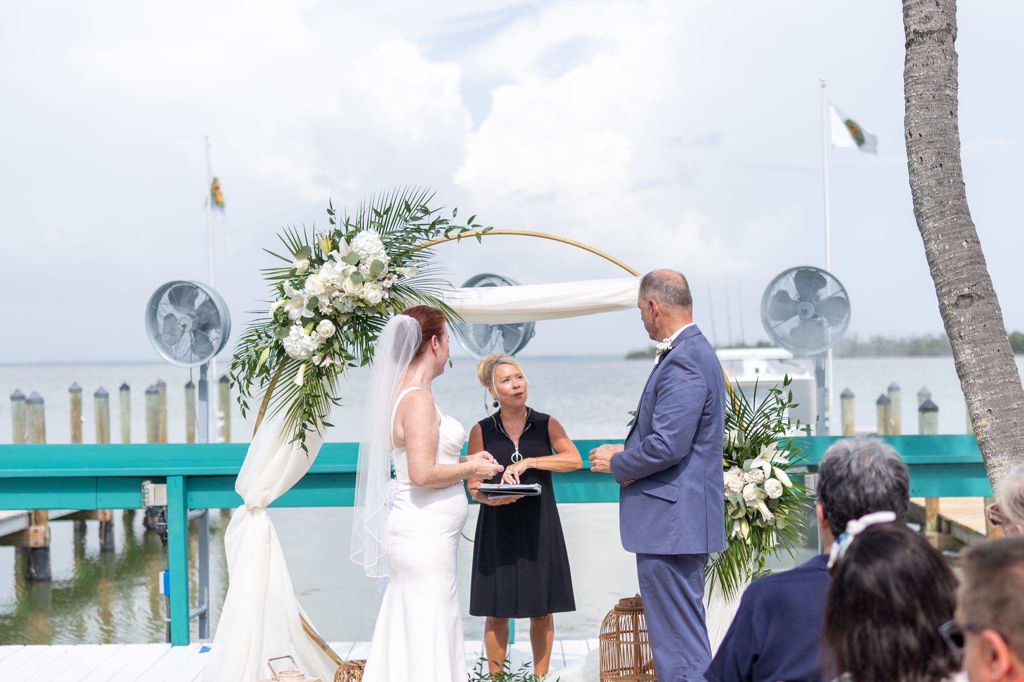 A bride and groom exchange vows on a waterfront dock beneath a circular floral arch as officiant Beth Schell leads the ceremony, palms and the marina behind them.