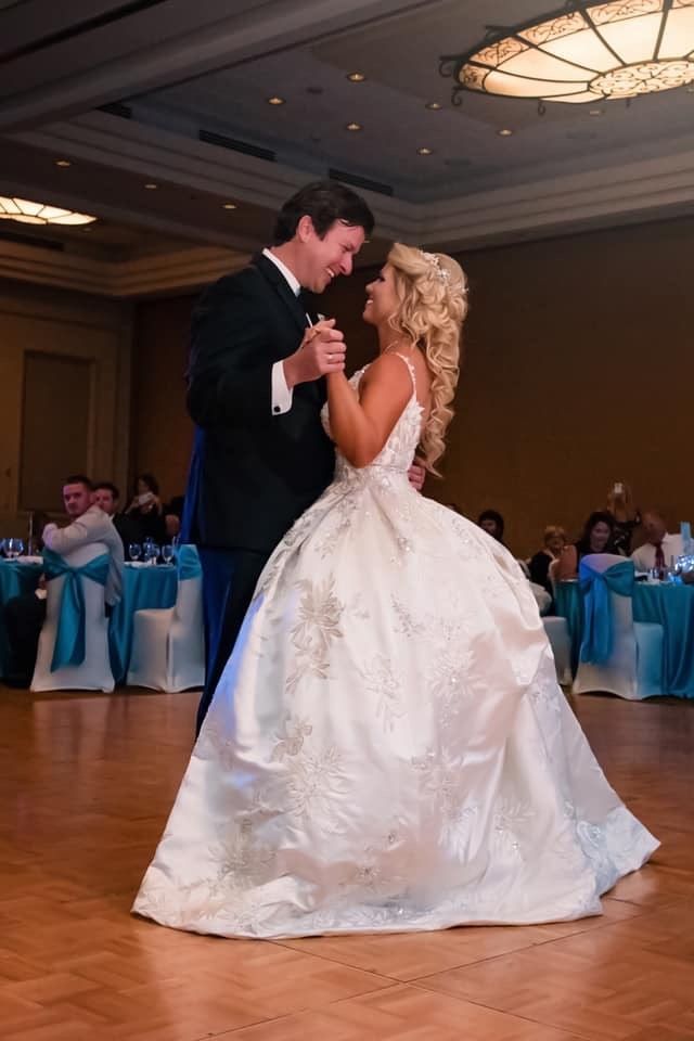 A bride in a beaded ball gown and her tuxedoed groom share their first dance in a warmly lit ballroom.