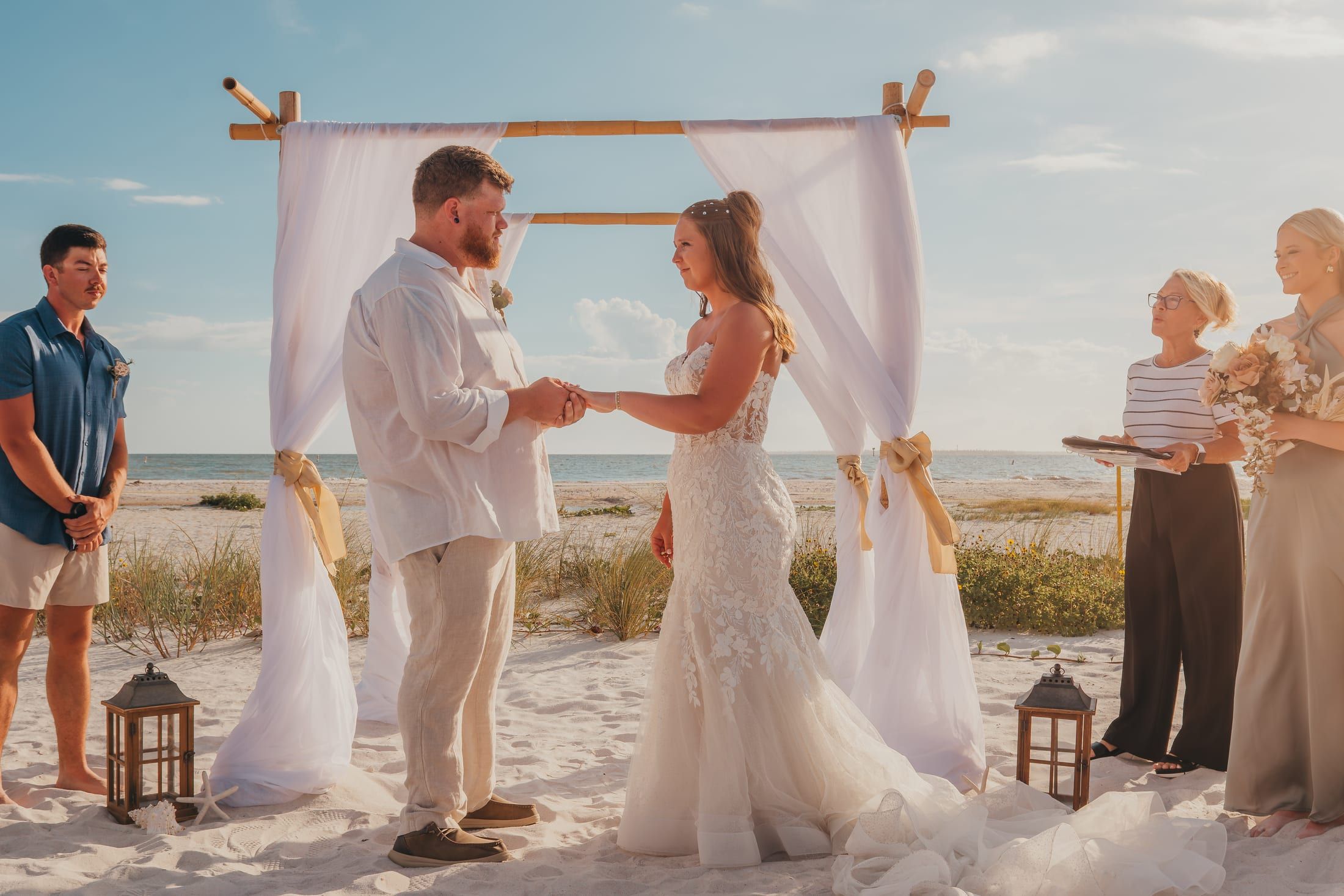 A bride and groom exchange rings at golden hour under a white draped beach arch while officiant Beth Schell reads the ceremony.