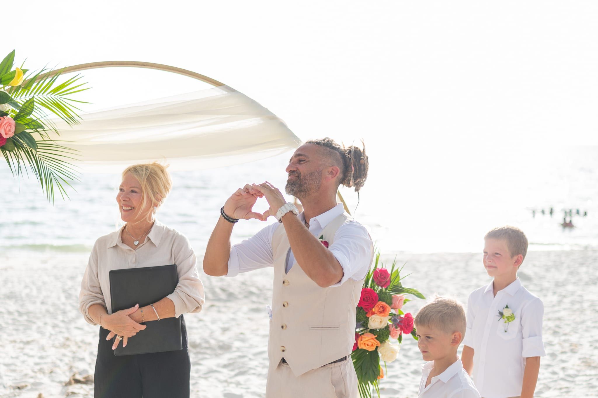 A joyful groom makes a heart with his hands as officiant Beth Schell beams beside him under the beach arch.
