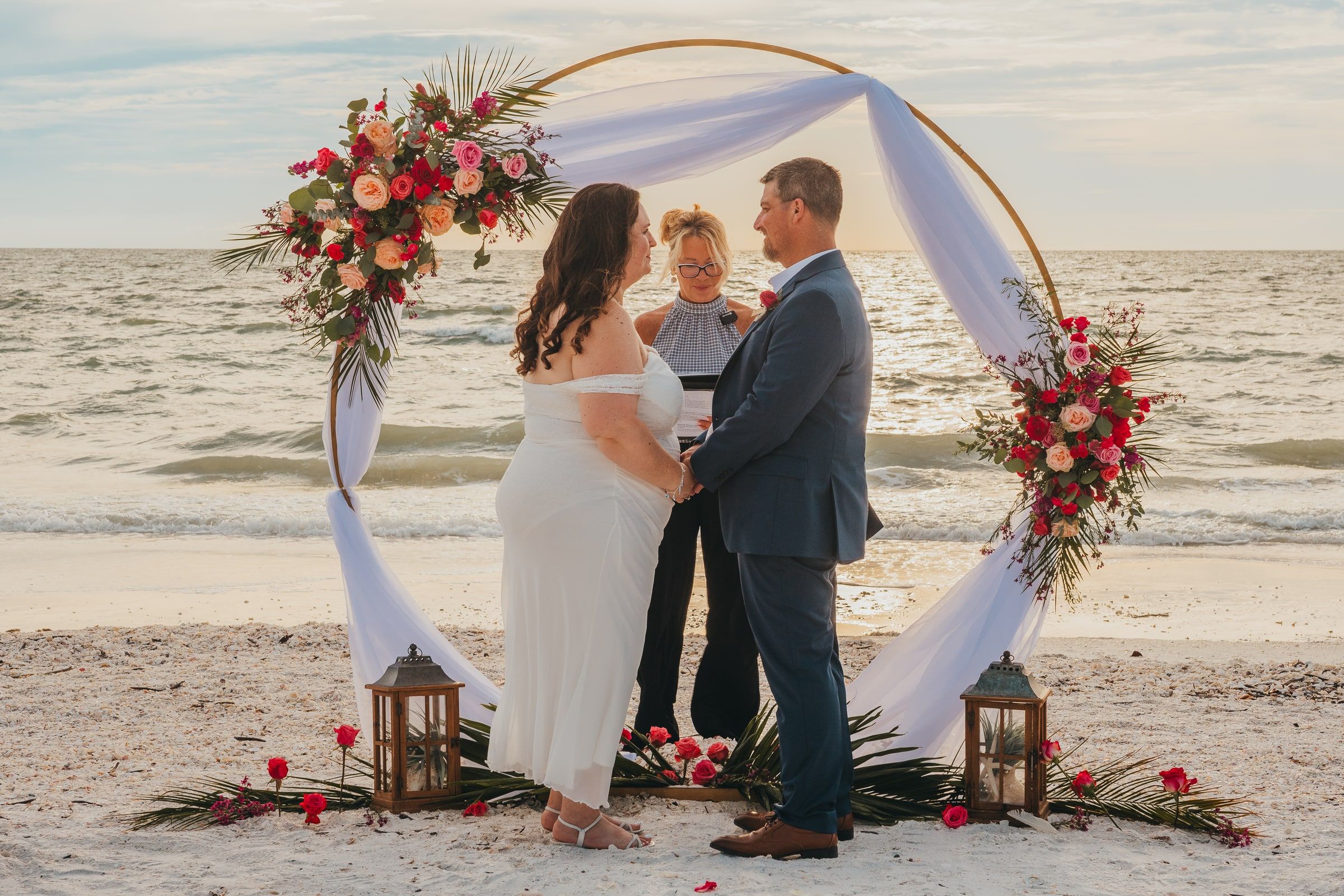A bride and groom hold hands under a golden floral arch at sunset while officiant Beth Schell leads their ceremony.