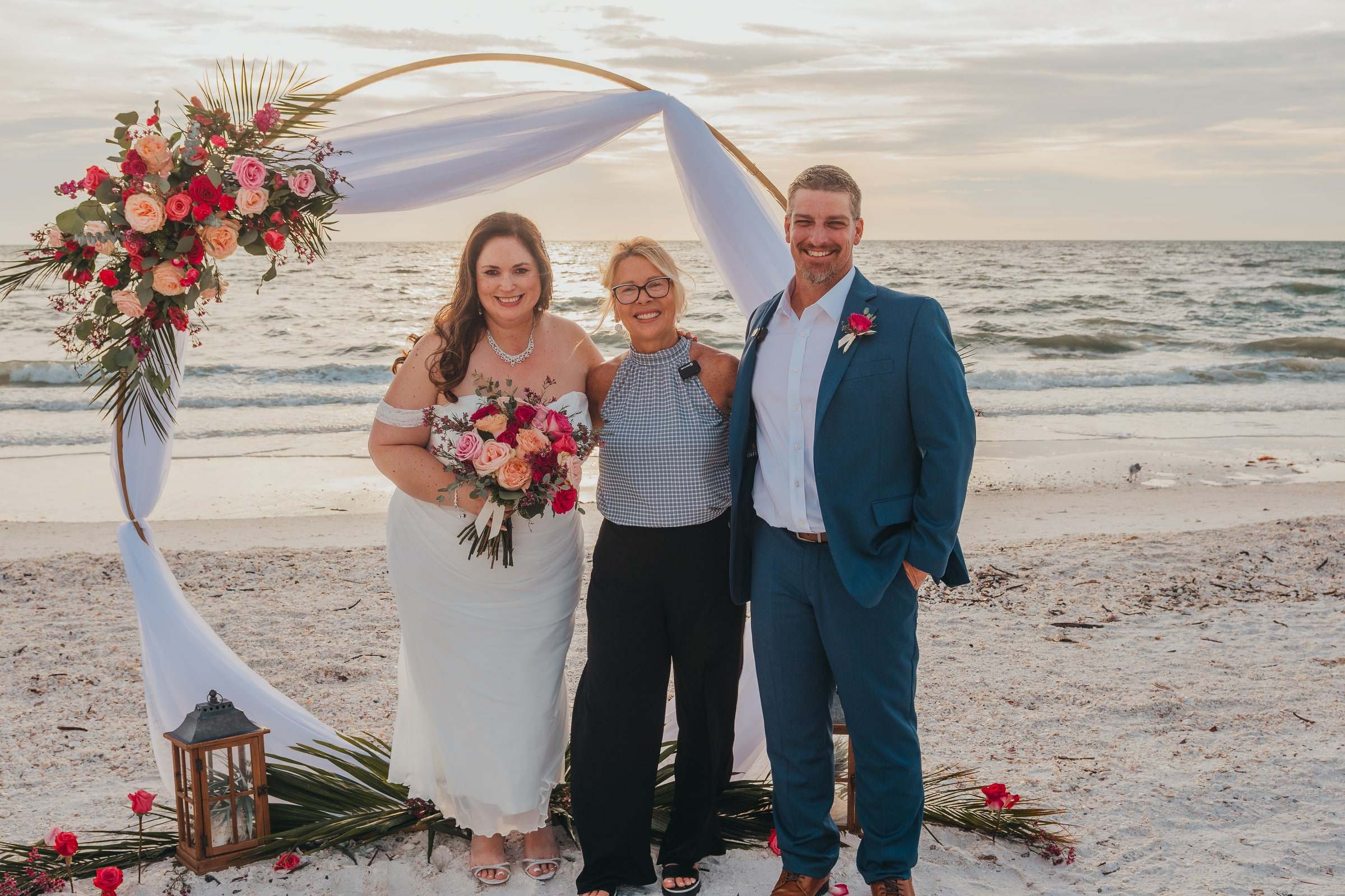 Officiant Beth Schell stands smiling between a bride and groom beneath a circular floral arch on the beach at sunset, the Gulf of Mexico behind them.