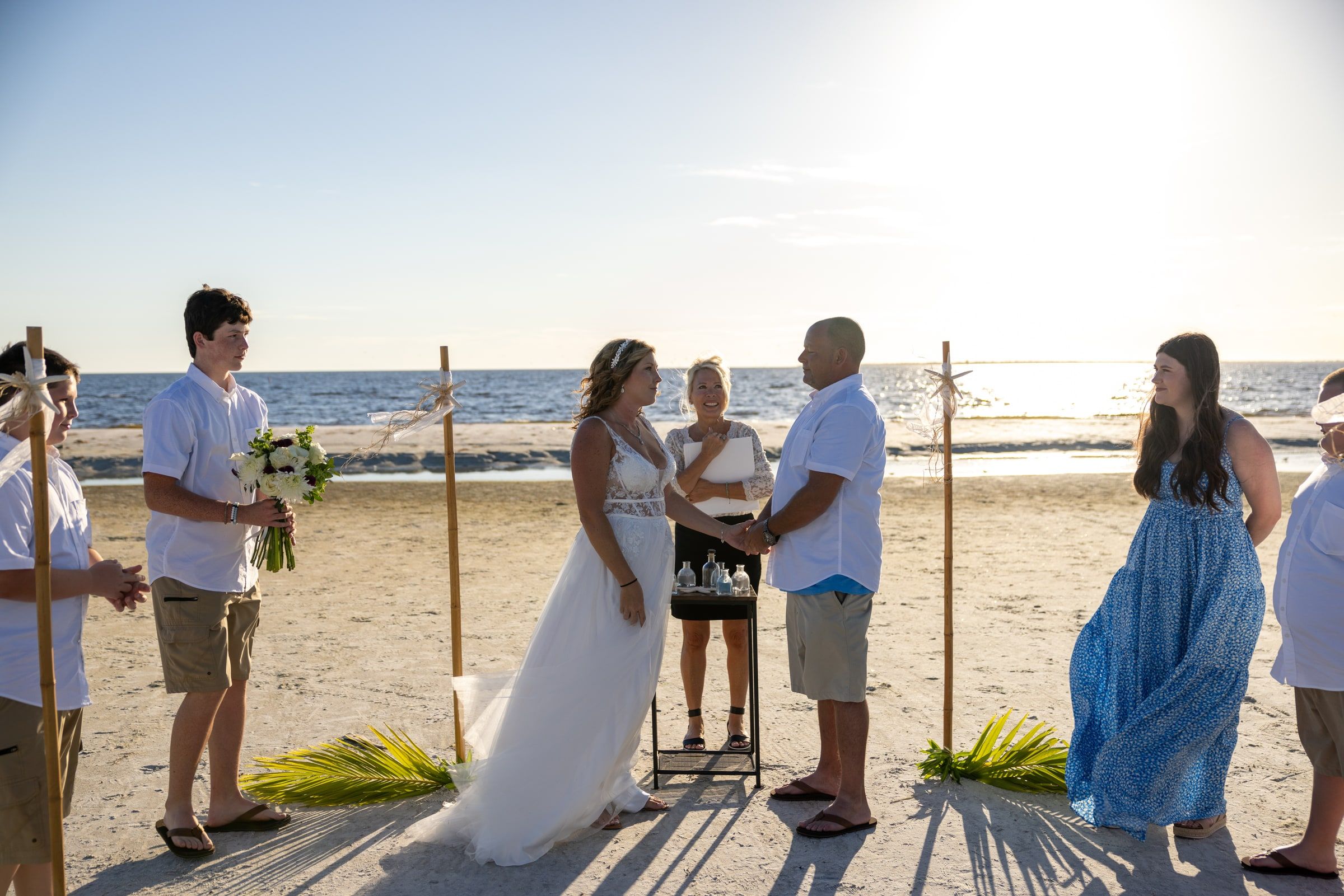 A couple holds hands during their beach wedding as officiant Beth Schell smiles between them, family gathered on both sides and the sun setting over the sea.
