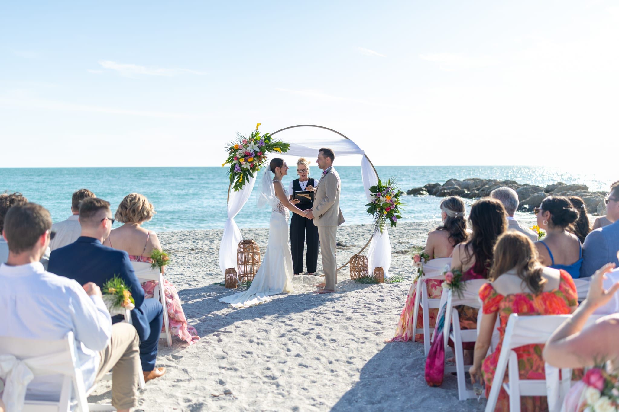 A bride and groom stand before a circular floral arch on the beach as officiant Beth Schell leads their vows, guests seated nearby.