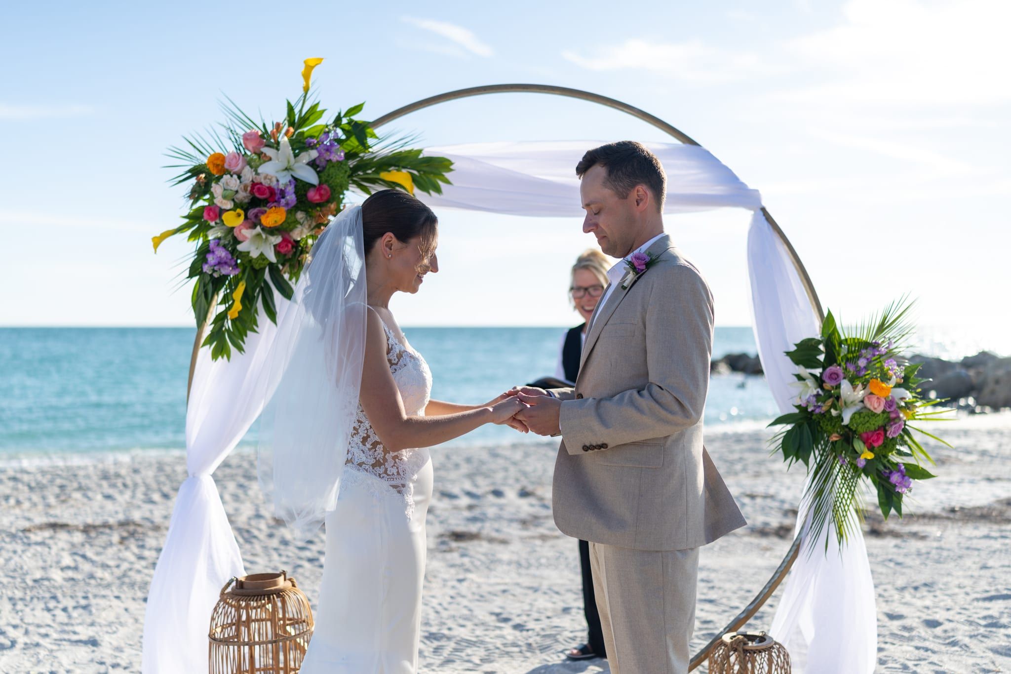 A couple exchanges rings before a colorful circular floral arch on the beach as officiant Beth Schell smiles beside them.
