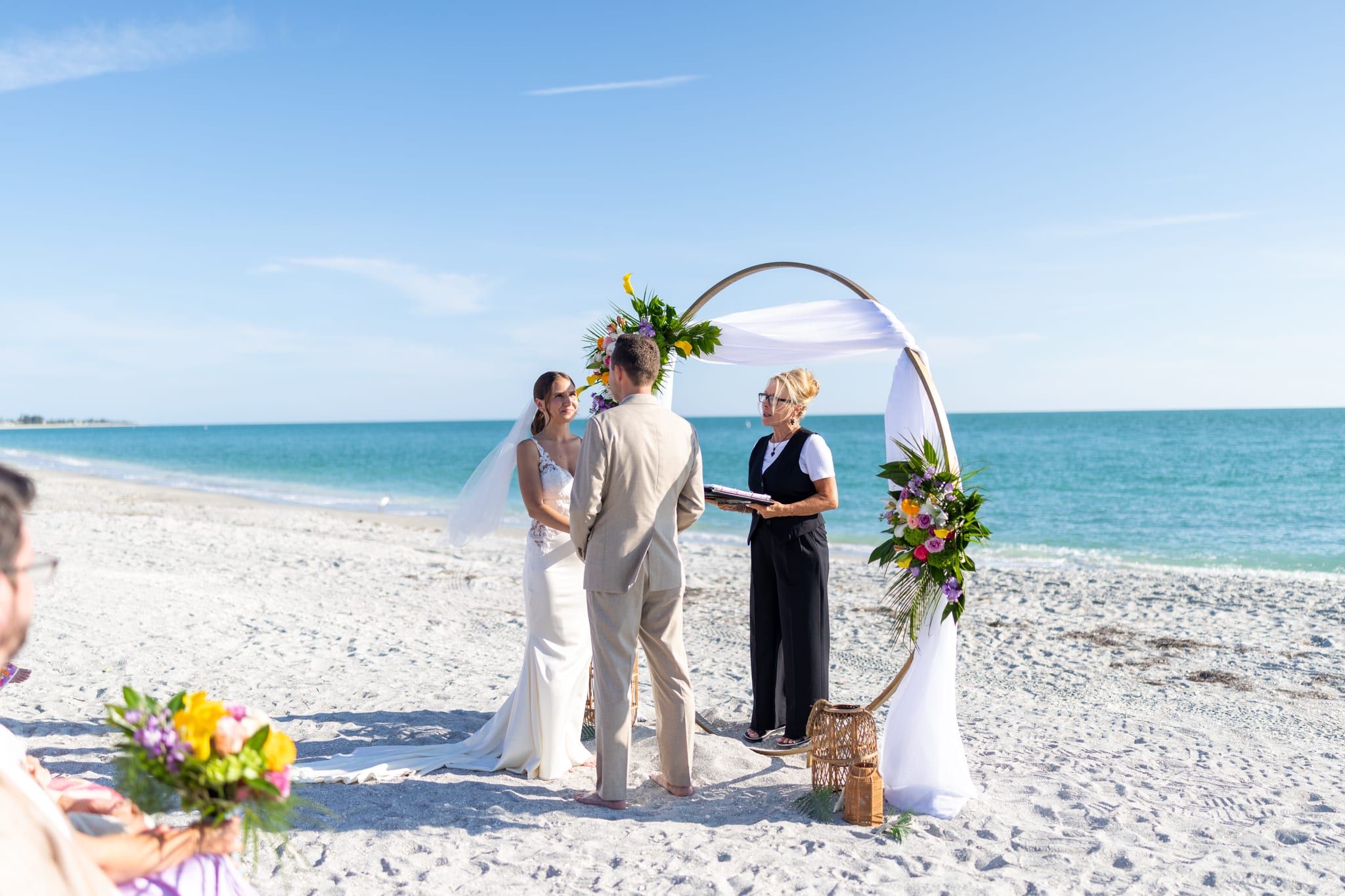 A bride and groom hold hands before a circular floral arch beside the turquoise Gulf of Mexico as officiant Beth Schell leads their ceremony.