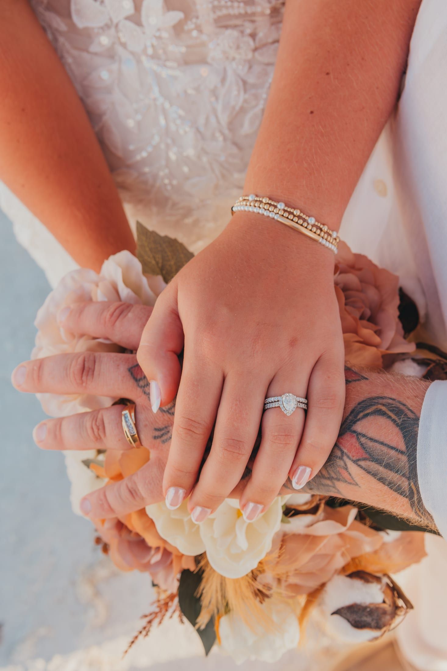 A close view of a couple’s joined hands resting on a boho bouquet, the pear-shaped engagement ring and wedding band catching the light.