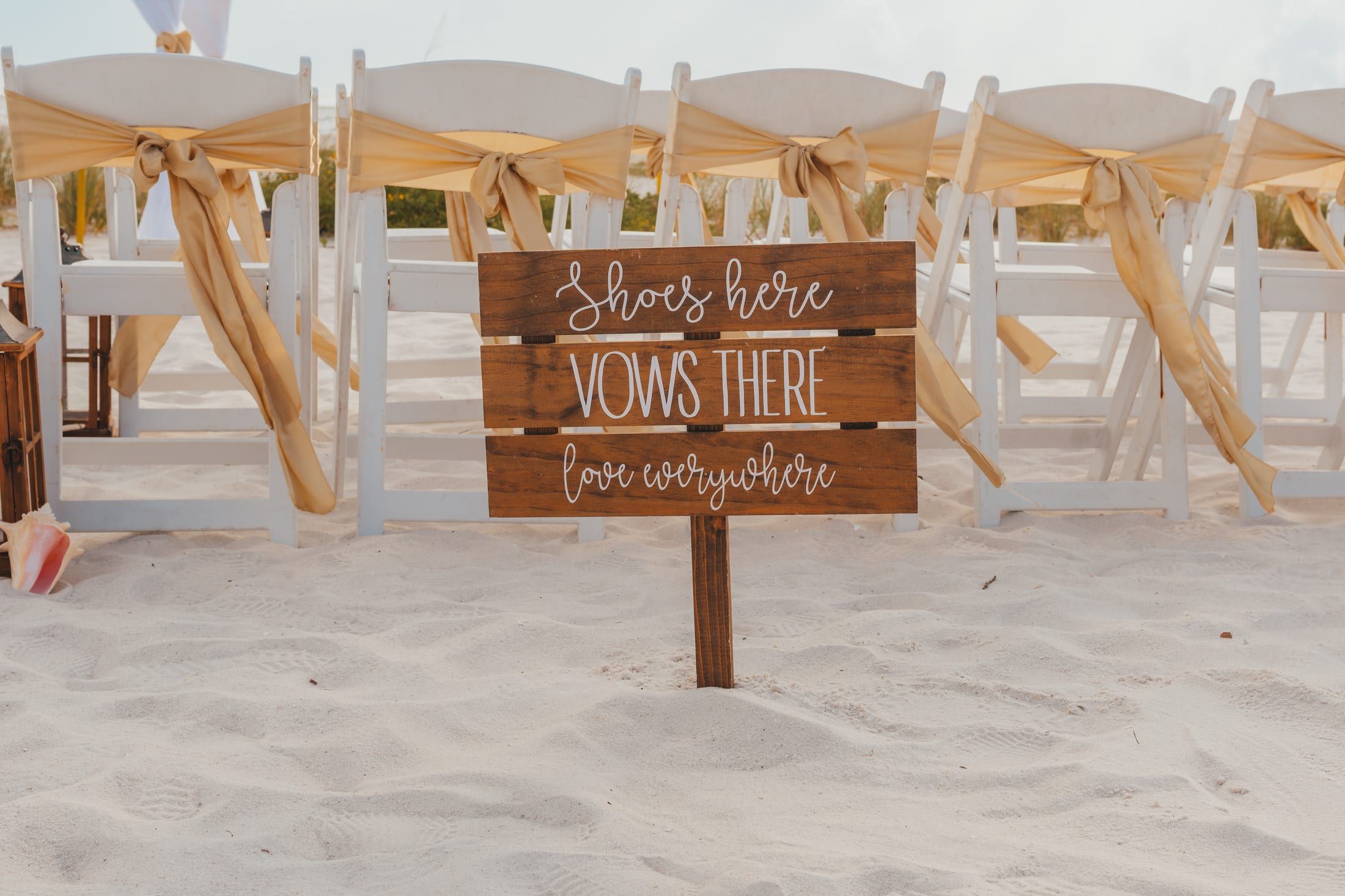 A wooden beach sign reading Shoes here, Vows there, love everywhere stands before white folding chairs tied with gold sashes on the sand.