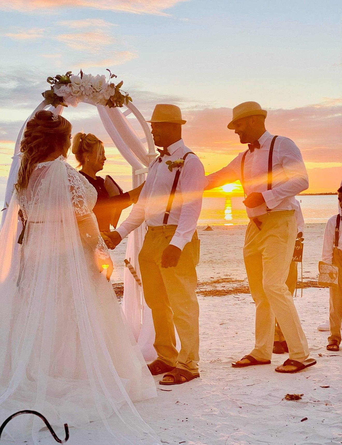 A bride and groom hold hands under a floral arch at sunset as officiant Beth Schell leads their beachside ceremony.