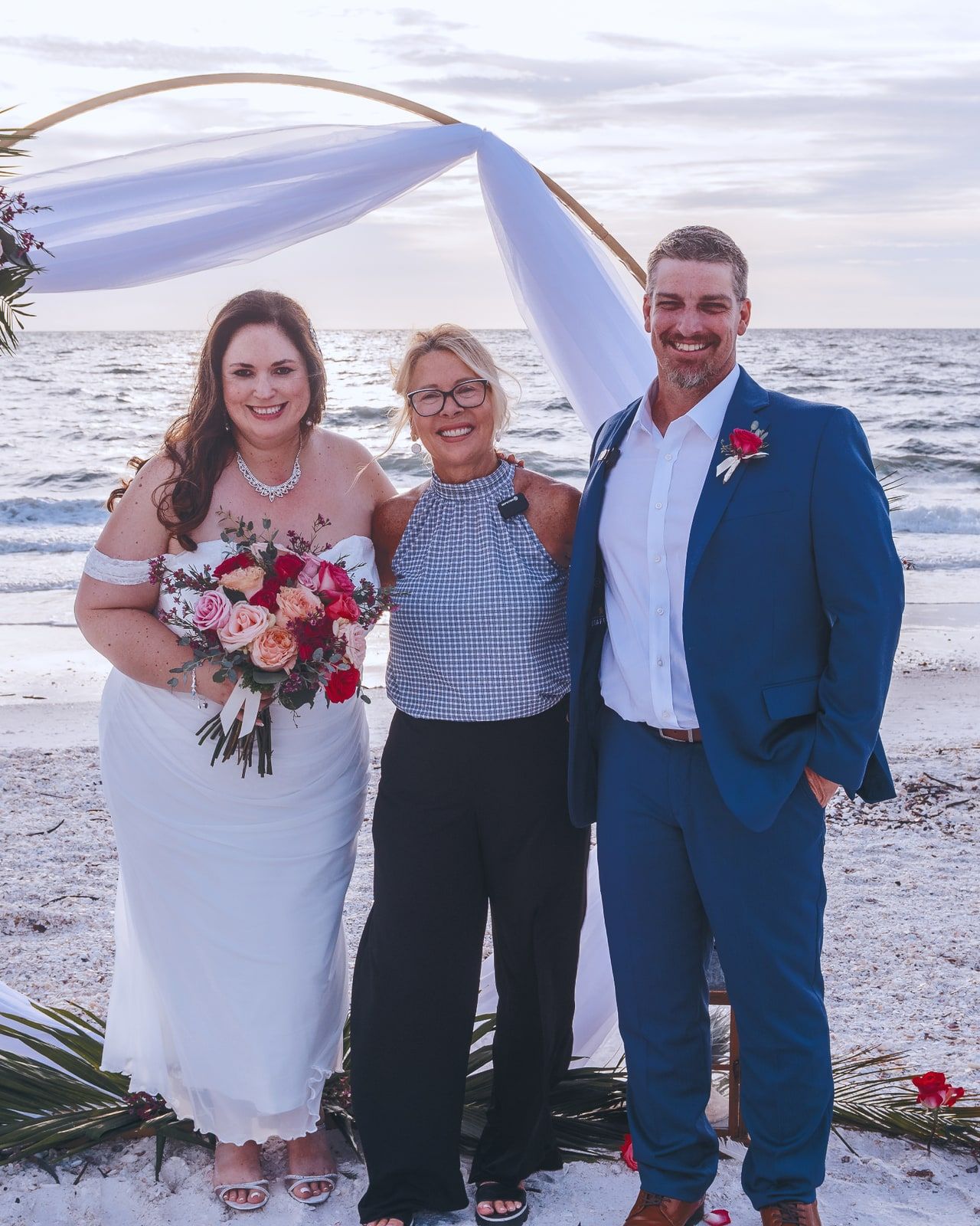 A newlywed couple stands smiling with officiant Beth Schell beside a beachfront floral arch, the bride holding a bouquet of pink and coral roses.