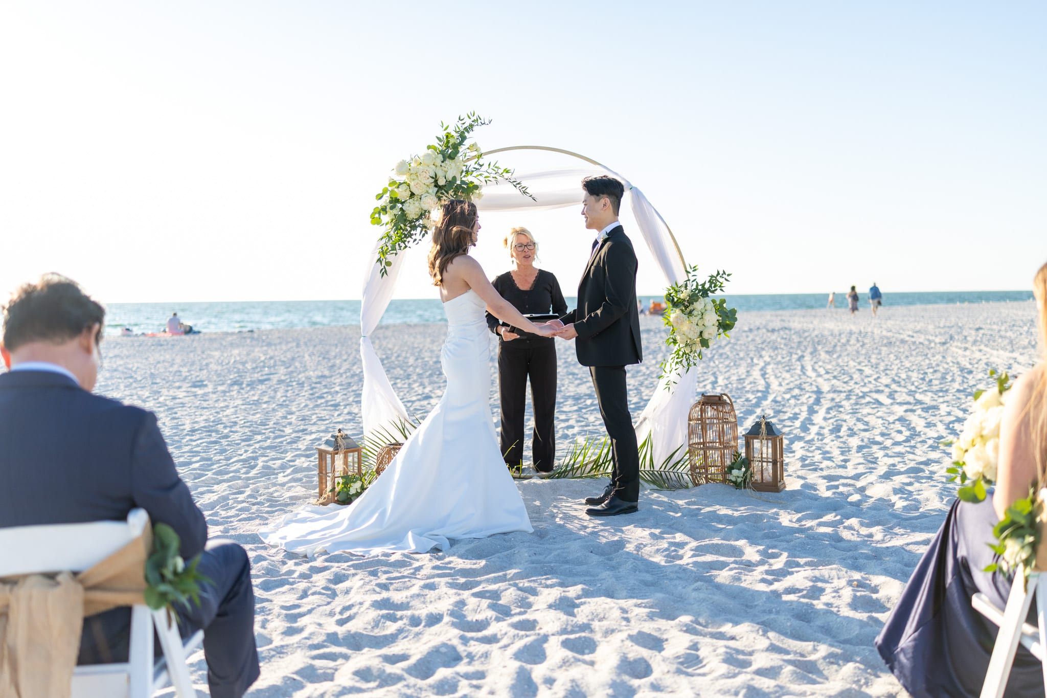 A bride and groom hold hands before a circular white floral arch on the beach as officiant Beth Schell officiates.